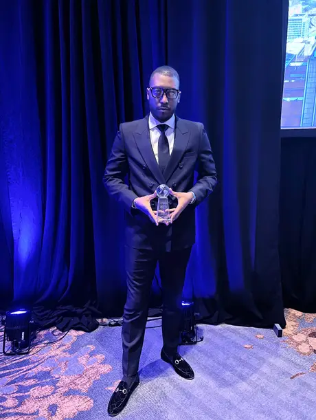Aton holding a crystal award beside blue stage curtains